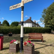 A vintage road sign with flower boxes and a bench on a grassy area under a blue sky.