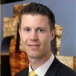 A man in a suit and tie smiles at the camera with a blurred artistic background behind him.
