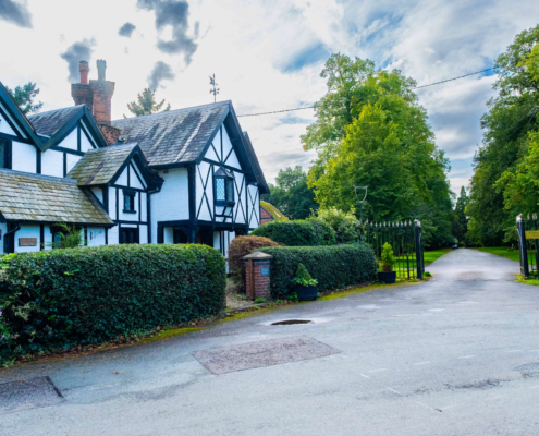 A white Tudor-style house with black trim beside a gated driveway lined with tall green trees.
