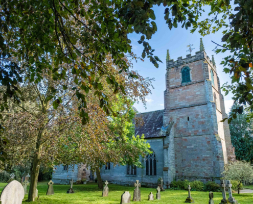 Stone church with a square tower surrounded by gravestones and trees on a sunny day.