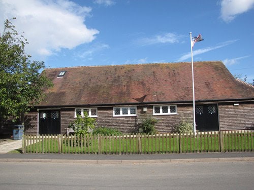 A wooden building with a red roof, a flagpole, and a small fenced lawn under a partly cloudy sky.