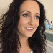 Woman with long curly dark hair, blue eyes, and a white top, smiling in a softly lit indoor setting.